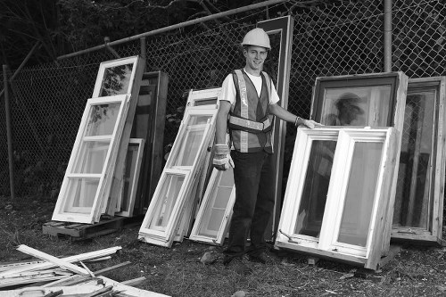 A man wearing a white hard hat, high-visibility vest, and work clothes is standing outdoors on a grassy area used for rubbish collection in Bow. He is smiling and holding a large, white window frame with a glass pane, positioned in front of several similar window frames that are leaning upright against a metal fence with a chain-link pattern. The frames are made of white painted wood or UPVC, featuring multiple glass panels and decorative edges, and are arranged in a line on the ground, with some resting directly on wooden pallets. The background includes trees and the fence, indicating a residential or commercial setting, with natural daylight illuminating the scene. The image relates to rubbish removal services provided by Garden Clearance Bow, focusing on the collection and disposal of discarded window frames and similar waste materials in the Bow area, suggesting a professional and organized approach to garden and property clearance.