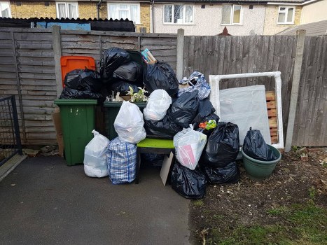 A collection of discarded rubbish including black bin bags, white plastic bags, and a checkered bag piled on a small wooden bench and surrounding area in a residential outdoor space. The rubbish is stacked against a wooden fence with weathered gray panels, behind which a green outdoor waste bin and an orange lid container are visible. To the right, a white metal bed frame or headboard is leaning against the fence, next to a large, empty green plastic plant container. The ground is paved with concrete, and there is a small patch of soil or dirt to the far right. This scene appears to be part of a garden clearance or rubbish removal process at a property in Bow, London, and is indicative of general waste collection efforts typical of local rubbish removal services like Garden Clearance Bow.