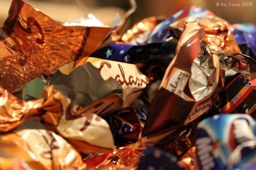 A close-up image showing a pile of crumpled foil and plastic wrappers in various metallic, matte, and glossy finishes, primarily in copper, silver, and colorful printed designs. The wrappers appear tangled and layered, with some edges curled and others overlapping, creating a textured and chaotic visual. The background is blurred, suggesting an indoor setting, possibly on a surface like a kitchen countertop or table. This collection of discarded packaging relates to rubbish and waste disposal, typical of household waste removal services offered by companies like Garden Clearance Bow. The focus is on the detailed textures of the crumpled materials, emphasizing their reflective surfaces and vibrant colors, with natural lighting highlighting the metallic sheen and creases in the wrappers.