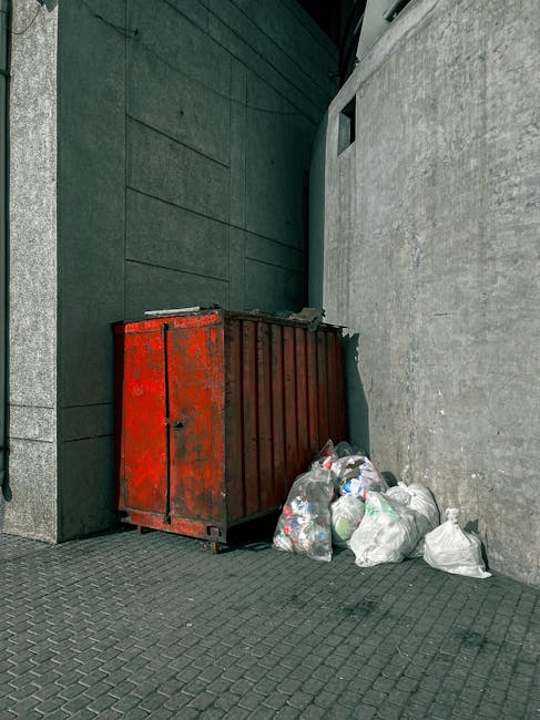 A red metal skip with a weathered, chipped finish is positioned against a dark green wall on the left and a light grey concrete wall on the right, situated outdoors on a textured grey paving surface. 
