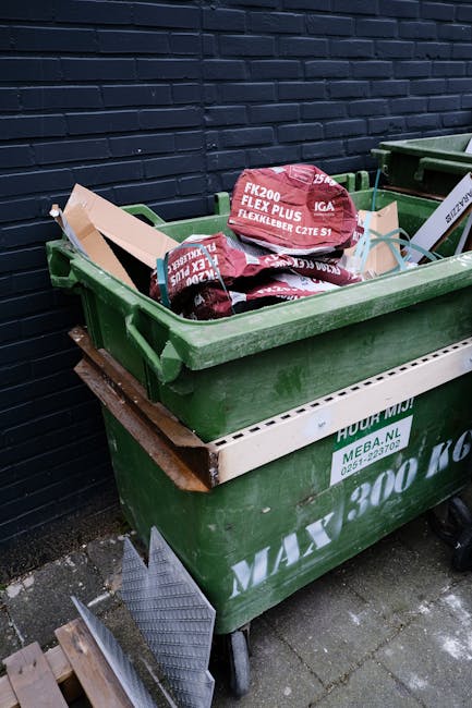 A red metal skip with a weathered, chipped finish is positioned against a dark green wall on the left and a light grey concrete wall on the right, situated outdoors on a textured grey paving surface. 