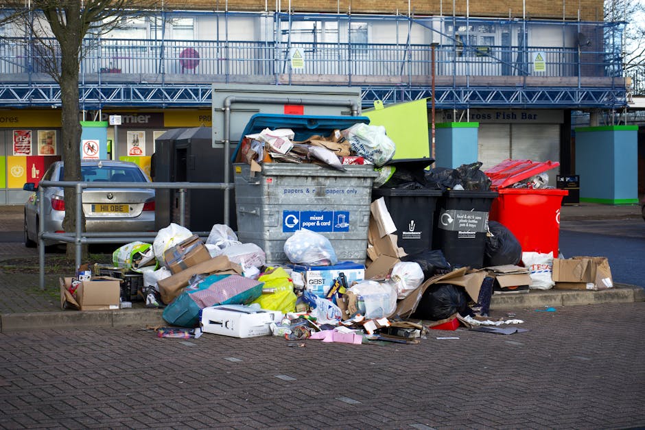 A large pile of mixed waste, including paper, cardboard boxes, plastic bags, and packaging materials, is overflowing from a grey recycling container labeled for mixed paper and cardboard, situated on