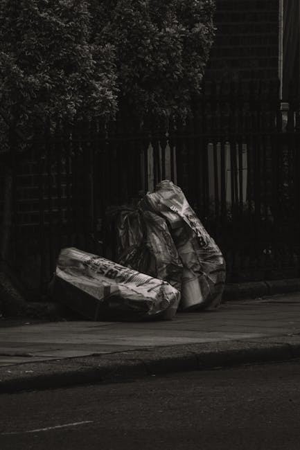 A large pile of mixed waste, including paper, cardboard boxes, plastic bags, and packaging materials, is overflowing from a grey recycling container labeled for mixed paper and cardboard, situated on