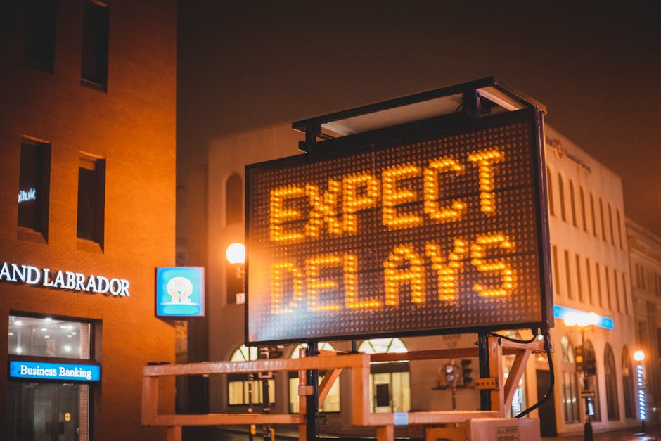 A large illuminated electronic road sign displaying the message 'EXPECT DELAYS' in bright orange LED lights is positioned on a street at night. The sign is mounted on a metal support framework and sta